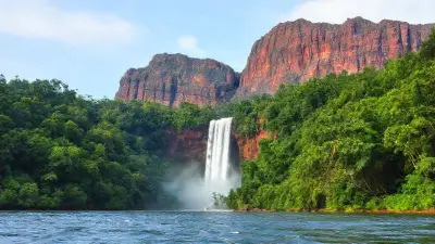 A dense, lush tropical forest is seen in the foreground. The trees are dense and vibrant green tones.

In front of the forest is a flowing river or lake shore. The water surface is slightly wavy.

A mountain of huge, dark reddish-brown sheer blocks of rock rises in the background.

A thin and very high waterfall flows down from the top of the mountain. The long and thin structure of Angel Falls is clearly evident.

White mist and clouds covered the lower part of the waterfall; This adds a mystical atmosphere to the landscape.

The sky is light blue, with some light clouds.
