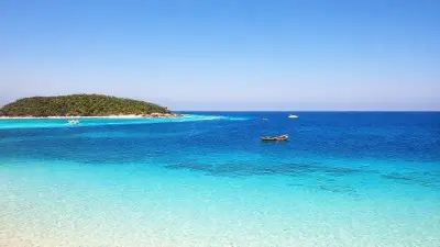 In the foreground lies an exceptionally clear ocean, transitioning from turquoise to deep blue.
The sea is so clear that even shallow patches of sand and rocky structures can be seen through the water.
The shoreline, shaped like a peninsula/ledge with a thin line of white sand extending into the sea, has created a natural beach language.

Beach and Land
The beach is covered with pristine white, soft, fine-grained sand.
Several wooden fishing boats and boats of various sizes are moored near the shore.
As you move inland, a densely verdant, wooded area opens up. Tropical trees and shrubs add a natural richness to the view.
Behind the trees, a more inland settlement, small structures, and a few larger buildings can be discerned.

Sea Movement
Across the vast ocean:

Several small boats,
Motorboats leaving a light trail on the water,
A small sailboat-like canoe-like vessel is seen moving in the wind on the left.

Atmosphere and Light
A time when the sun is nearing sunset or sunrise; slightly soft, golden yellow tones.
The sky is a light pastel shade, cloudless or slightly overcast.
The overall photo conveys the calm, warm, and peaceful feeling of a tropical island vacation.